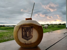 A coconut with a straw inserted sits on a wooden surface. The coconut is branded with the words 'Empire Fit Club Bali'. In the background, there is a lush green field and an overcast sky with orange-yellow tinges from a setting or rising sun.
