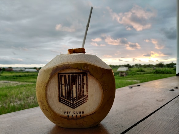 A coconut with a straw inserted sits on a wooden surface. The coconut is branded with the words 'Empire Fit Club Bali'. In the background, there is a lush green field and an overcast sky with orange-yellow tinges from a setting or rising sun.