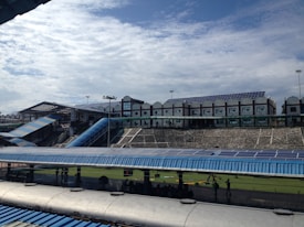 A large structure with numerous solar panels on the roof is visible, suggesting an eco-friendly or energy-efficient building. The building has a modern design with multiple windows and a blue color scheme. A blue, sloped walkway or canopy is present, leading from one section to another. The surrounding area includes green grass and some trees, with a bright, partially cloudy sky overhead.