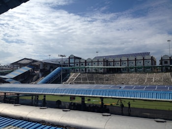 A large structure with numerous solar panels on the roof is visible, suggesting an eco-friendly or energy-efficient building. The building has a modern design with multiple windows and a blue color scheme. A blue, sloped walkway or canopy is present, leading from one section to another. The surrounding area includes green grass and some trees, with a bright, partially cloudy sky overhead.