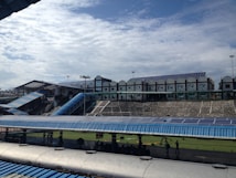 A large structure with numerous solar panels on the roof is visible, suggesting an eco-friendly or energy-efficient building. The building has a modern design with multiple windows and a blue color scheme. A blue, sloped walkway or canopy is present, leading from one section to another. The surrounding area includes green grass and some trees, with a bright, partially cloudy sky overhead.