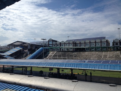 A large structure with numerous solar panels on the roof is visible, suggesting an eco-friendly or energy-efficient building. The building has a modern design with multiple windows and a blue color scheme. A blue, sloped walkway or canopy is present, leading from one section to another. The surrounding area includes green grass and some trees, with a bright, partially cloudy sky overhead.