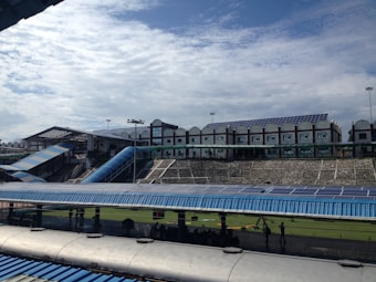 A large structure with numerous solar panels on the roof is visible, suggesting an eco-friendly or energy-efficient building. The building has a modern design with multiple windows and a blue color scheme. A blue, sloped walkway or canopy is present, leading from one section to another. The surrounding area includes green grass and some trees, with a bright, partially cloudy sky overhead.