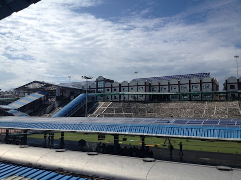 A large structure with numerous solar panels on the roof is visible, suggesting an eco-friendly or energy-efficient building. The building has a modern design with multiple windows and a blue color scheme. A blue, sloped walkway or canopy is present, leading from one section to another. The surrounding area includes green grass and some trees, with a bright, partially cloudy sky overhead.