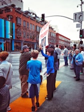 A group of people standing on a city street corner, with several holding a large sign that reads 'Stop Trump'. The scene is set in an urban environment with brick and glass buildings. The atmosphere appears busy, with pedestrians waiting on the sidewalk, some interacting with each other. A traffic light and road signs are visible in the background.