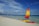 A colorful sailboat with a vibrant, multi-colored sail is stationed on the sandy beach next to a red kayak. The clear blue sea extends to the horizon with several people wading and swimming in the shallow waters. The sky is mostly clear with some wispy clouds. A few boats are visible in the distance, and there are people walking along the shoreline. The overall scenery is of a peaceful beach setting with clear skies and calm waters.