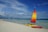 A colorful sailboat with a vibrant, multi-colored sail is stationed on the sandy beach next to a red kayak. The clear blue sea extends to the horizon with several people wading and swimming in the shallow waters. The sky is mostly clear with some wispy clouds. A few boats are visible in the distance, and there are people walking along the shoreline. The overall scenery is of a peaceful beach setting with clear skies and calm waters.