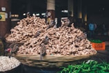 A bustling market scene featuring a large pile of ginger in a woven basket with several small birds perched on the ginger. Surrounding the ginger are piles of other produce such as garlic and green chilies in crates. The market is dimly lit, with people visible in the background.