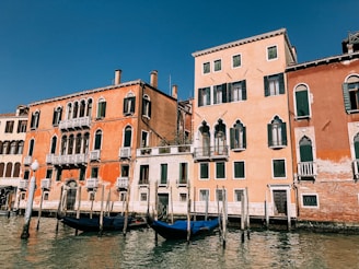 Traditional Venetian buildings with colorful facades line a canal. The architecture features arched windows and small balconies with ornate railings. Two gondolas are docked at the water's edge, reflecting on the canal's surface.