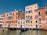 Traditional Venetian buildings with colorful facades line a canal. The architecture features arched windows and small balconies with ornate railings. Two gondolas are docked at the water's edge, reflecting on the canal's surface.
