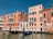 Traditional Venetian buildings with colorful facades line a canal. The architecture features arched windows and small balconies with ornate railings. Two gondolas are docked at the water's edge, reflecting on the canal's surface.