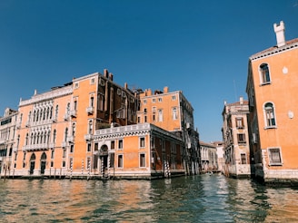Renaissance-style orange buildings line both sides of a canal under a clear blue sky. The water in the canal is calm, reflecting the architecture. Ornate windows, arched doorways, and balconies add charm to the scene.