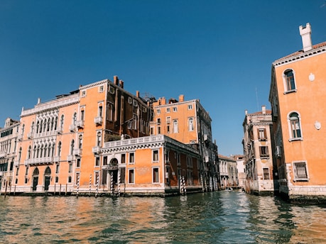 Renaissance-style orange buildings line both sides of a canal under a clear blue sky. The water in the canal is calm, reflecting the architecture. Ornate windows, arched doorways, and balconies add charm to the scene.