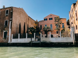 A picturesque view of colorful Venetian buildings facing a canal. The architecture features arched windows, rustic brickwork, and vibrant facades. Tall, slender trees and palm trees are visible near the water's edge, complementing the serene atmosphere.