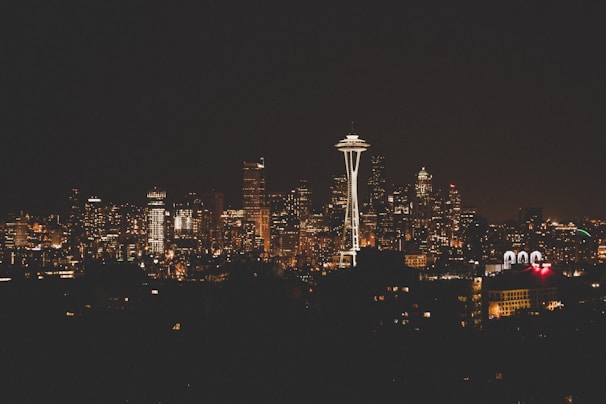 The city skyline at night is illuminated by numerous lights from the buildings, with the iconic Space Needle prominently standing out against the dark sky.