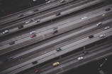 An aerial view of a multi-lane highway with several vehicles traveling in both directions. The roads are elevated and parallel to each other, with a grayish tone dominating the scene. Vehicles of varying colors and sizes are visible, with noticeable space between them, suggesting moderate traffic.