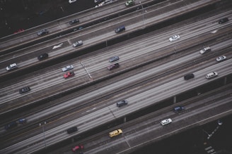 An aerial view of a multi-lane highway with several vehicles traveling in both directions. The roads are elevated and parallel to each other, with a grayish tone dominating the scene. Vehicles of varying colors and sizes are visible, with noticeable space between them, suggesting moderate traffic.