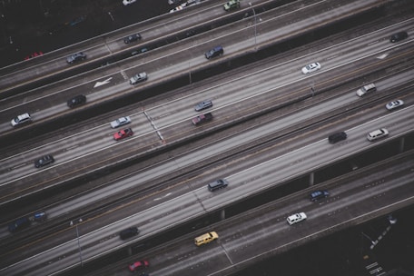 An aerial view of a multi-lane highway with several vehicles traveling in both directions. The roads are elevated and parallel to each other, with a grayish tone dominating the scene. Vehicles of varying colors and sizes are visible, with noticeable space between them, suggesting moderate traffic.