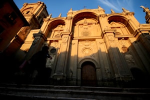 An ornate, historical facade of a cathedral bathed in warm, golden sunlight, casting long shadows. The intricate architectural details include columns, arches, and religious carvings, conveying a sense of grandeur and historical significance. A silhouette of a person walks in the foreground, highlighting the massive scale of the building.