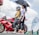 A motorcyclist wearing a helmet and protective gear sits on a red Kawasaki bike marked with the number 88. Next to the motorcyclist, a woman holds a black umbrella to provide shade. They are outdoors, and in the background, there is a flag with 'Mutual' branding and another person standing near similar banners. The sky is partly cloudy.
