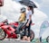 A motorcyclist wearing a helmet and protective gear sits on a red Kawasaki bike marked with the number 88. Next to the motorcyclist, a woman holds a black umbrella to provide shade. They are outdoors, and in the background, there is a flag with 'Mutual' branding and another person standing near similar banners. The sky is partly cloudy.