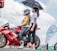 A motorcyclist wearing a helmet and protective gear sits on a red Kawasaki bike marked with the number 88. Next to the motorcyclist, a woman holds a black umbrella to provide shade. They are outdoors, and in the background, there is a flag with 'Mutual' branding and another person standing near similar banners. The sky is partly cloudy.