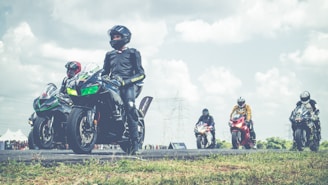 A group of motorcyclists in full gear are positioned on a racetrack, with several sport motorcycles in various colors. The sky is partly cloudy, and there are distant power lines visible. The setting appears to be an organized race or event, with a crowd in the background.