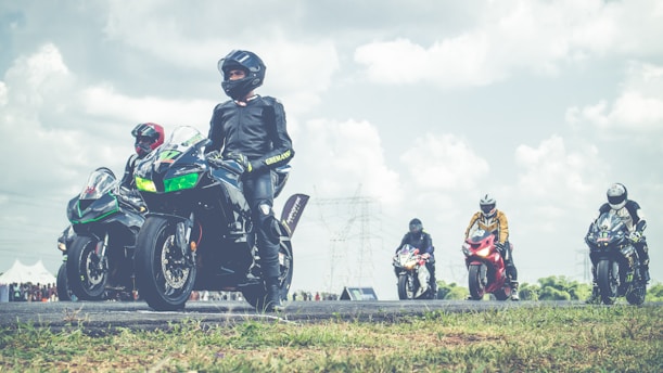A group of motorcyclists in full gear are positioned on a racetrack, with several sport motorcycles in various colors. The sky is partly cloudy, and there are distant power lines visible. The setting appears to be an organized race or event, with a crowd in the background.