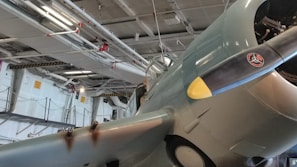 A vintage military aircraft is displayed indoors, partially elevated off the ground. The scene is filled with industrial elements, including pipes and overhead lights. The aircraft features distinctive markings on its propeller and faded paint, indicating its historic nature.