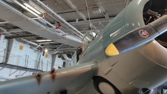 A vintage military aircraft is displayed indoors, partially elevated off the ground. The scene is filled with industrial elements, including pipes and overhead lights. The aircraft features distinctive markings on its propeller and faded paint, indicating its historic nature.