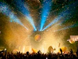 A vibrant concert or festival scene features a crowd of people with their hands raised, bright stage lighting, and a flurry of multicolored confetti filling the air. Large inflatable balls and decorations add to the festive atmosphere, with beams of blue and yellow light cutting through the space.