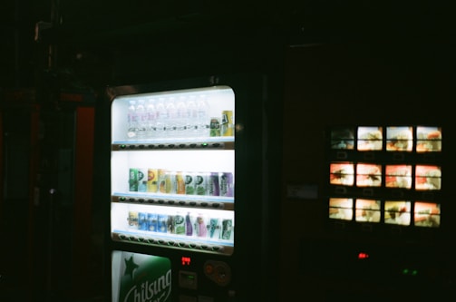 A dimly lit environment featuring a vending machine stocked with a variety of beverages, including water bottles and canned drinks, illuminated under a soft glow.