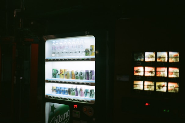 A dimly lit environment featuring a vending machine stocked with a variety of beverages, including water bottles and canned drinks, illuminated under a soft glow.