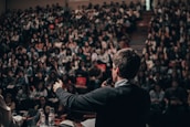 A large audience is seated in an auditorium, with a person standing at the front holding a microphone and addressing the crowd. The auditorium is filled with people, indicating an event or lecture taking place.