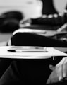 grayscale photo of students sitting on chairs with papers and pens