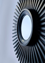A circular mirror with a black, sunburst-like frame composed of elongated, evenly spaced lines extending outward.