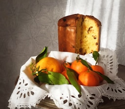 A traditional Italian panettone with a crusty, golden-brown exterior is displayed alongside fresh, vibrant oranges with green leaves. The items are placed on an intricately designed white cloth, casting soft shadows on a subtle textured background.