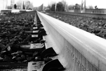 A detailed view of a railway track taken from a low angle. The track stretches into the distance, with gravel and railroad fittings clearly visible. The background is blurred, suggesting movement or depth. The image is in black and white, giving it a classic or timeless feel.
