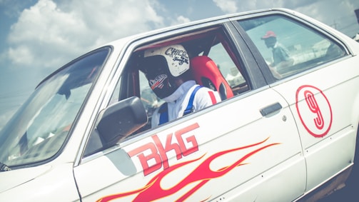 A person wearing a black and white helmet is seated inside a white sports car with racing decals, including red flames and the number 9. The words 'BKG' are prominently displayed on the side of the car. The car's window is down, and another person wearing a cap is visible faintly in the background.