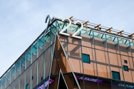 A modern building with glass facades and wooden paneling. The structure is topped with the large metallic numbers '222'. There are geometric patterns formed by the intersecting lines and panels, and a message scrolling in pink tint at the bottom.