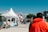 A busy outdoor market scene with numerous people walking and gathering around various stalls. The vendors are in white tents with blue text, offering food and other goods. The sky is clear and blue, suggesting good weather, while trees and buildings are visible in the background. The atmosphere feels lively with interactions among the crowd.
