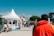 A busy outdoor market scene with numerous people walking and gathering around various stalls. The vendors are in white tents with blue text, offering food and other goods. The sky is clear and blue, suggesting good weather, while trees and buildings are visible in the background. The atmosphere feels lively with interactions among the crowd.