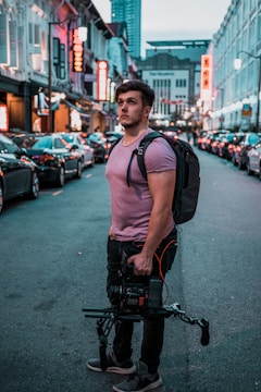 A person stands on a city street holding professional camera equipment. The street is lined with cars, and lit by neon signs and lights from shops and restaurants. The person is wearing a backpack and casual clothing, suggesting they may be preparing to film or take photographs. In the background, buildings and high-rises are visible under an early evening sky.