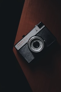 A moody, dimly lit photo of a vintage camera resting on a dark wooden table with a faint reddish glow.