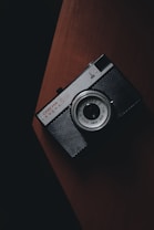 A vintage camera with a black body and silver lens sits on a wooden surface, captured with dim lighting that creates a moody and classic aesthetic.