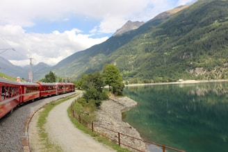 A red train travels along a curved railway track next to a serene body of water, surrounded by lush green hills and mountains under a partly cloudy sky. The scene is picturesque, with layers of nature contrasted by the vibrant color of the train.