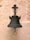 A vintage metal bell is mounted on a textured stone wall. The bell has a dark patina and is designed with a circular handle at its base. The mounting bracket features a cross-like structure with a rusted appearance.
