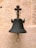 A vintage metal bell is mounted on a textured stone wall. The bell has a dark patina and is designed with a circular handle at its base. The mounting bracket features a cross-like structure with a rusted appearance.