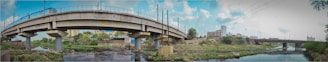 A panoramic view of two bridges crossing over a small body of water, with one bridge having a curved design supported by thick columns. The surrounding area includes sparse vegetation, construction debris, and buildings in the distance under a partly cloudy sky. Electrical poles and wires are visible along the bridges.