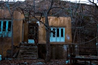 An abandoned house stands amid barren trees. The structure appears weathered, with peeling paint and broken windows. The steps leading to the entrance are cracked, and the surrounding ground is covered with dry leaves. The atmosphere is desolate and haunting.